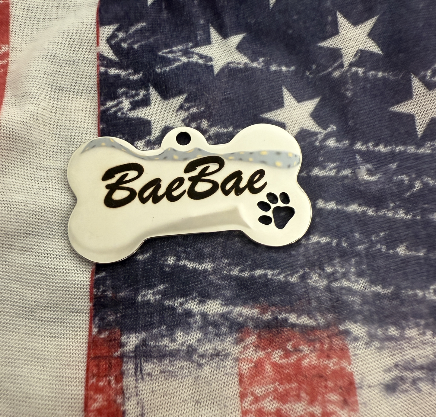 Personalized stainless steel pet ID tag shaped like a bone with custom engraving "BaeBae" and a paw print on an American flag background