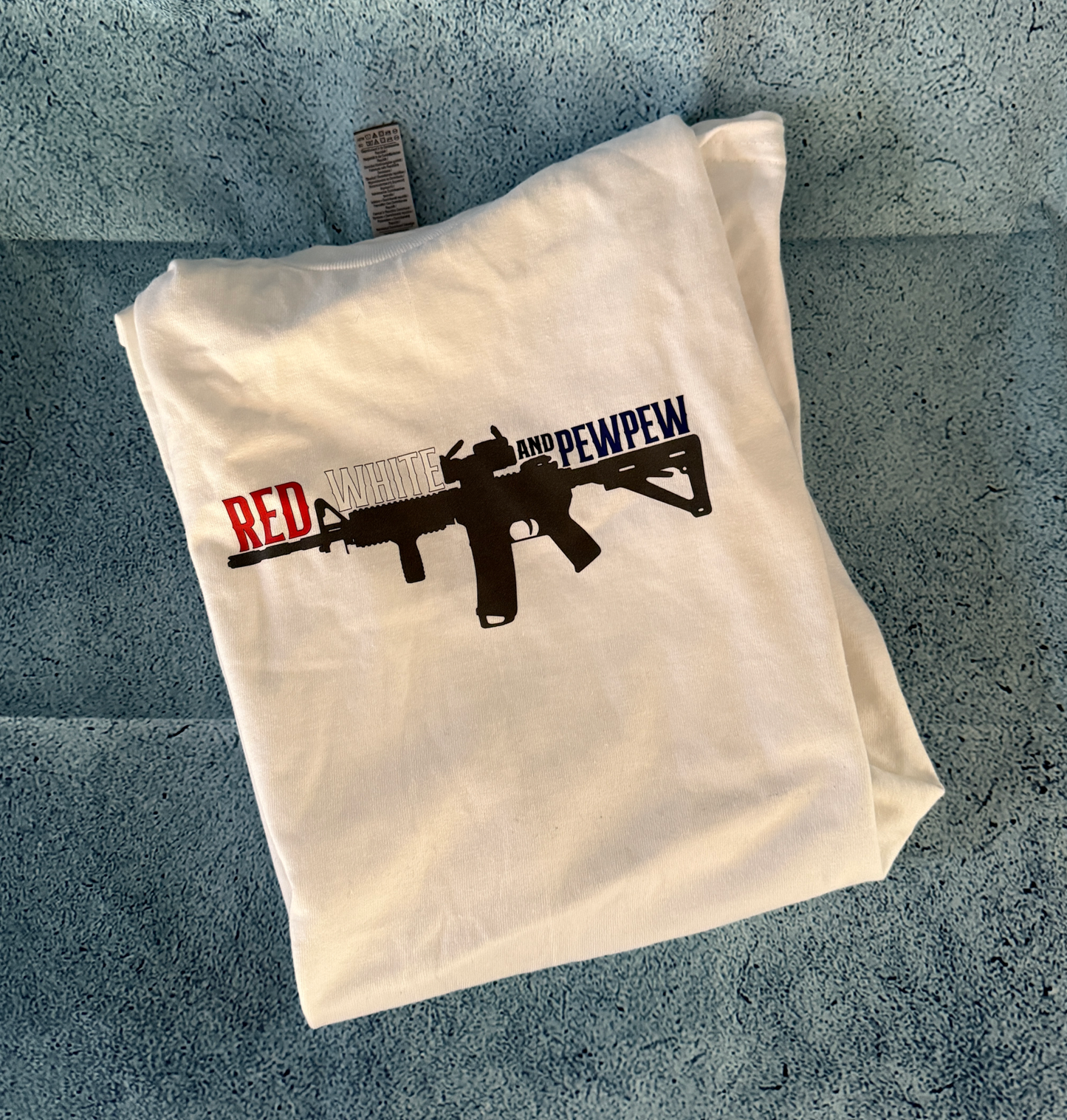 White folded t-shirt with "Red White and PewPew" text and black rifle graphic design