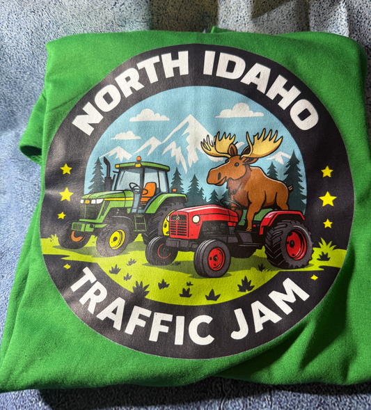 Green t-shirt with a graphic of tractors and a moose, labeled 'North Idaho Traffic Jam', on a textured surface.