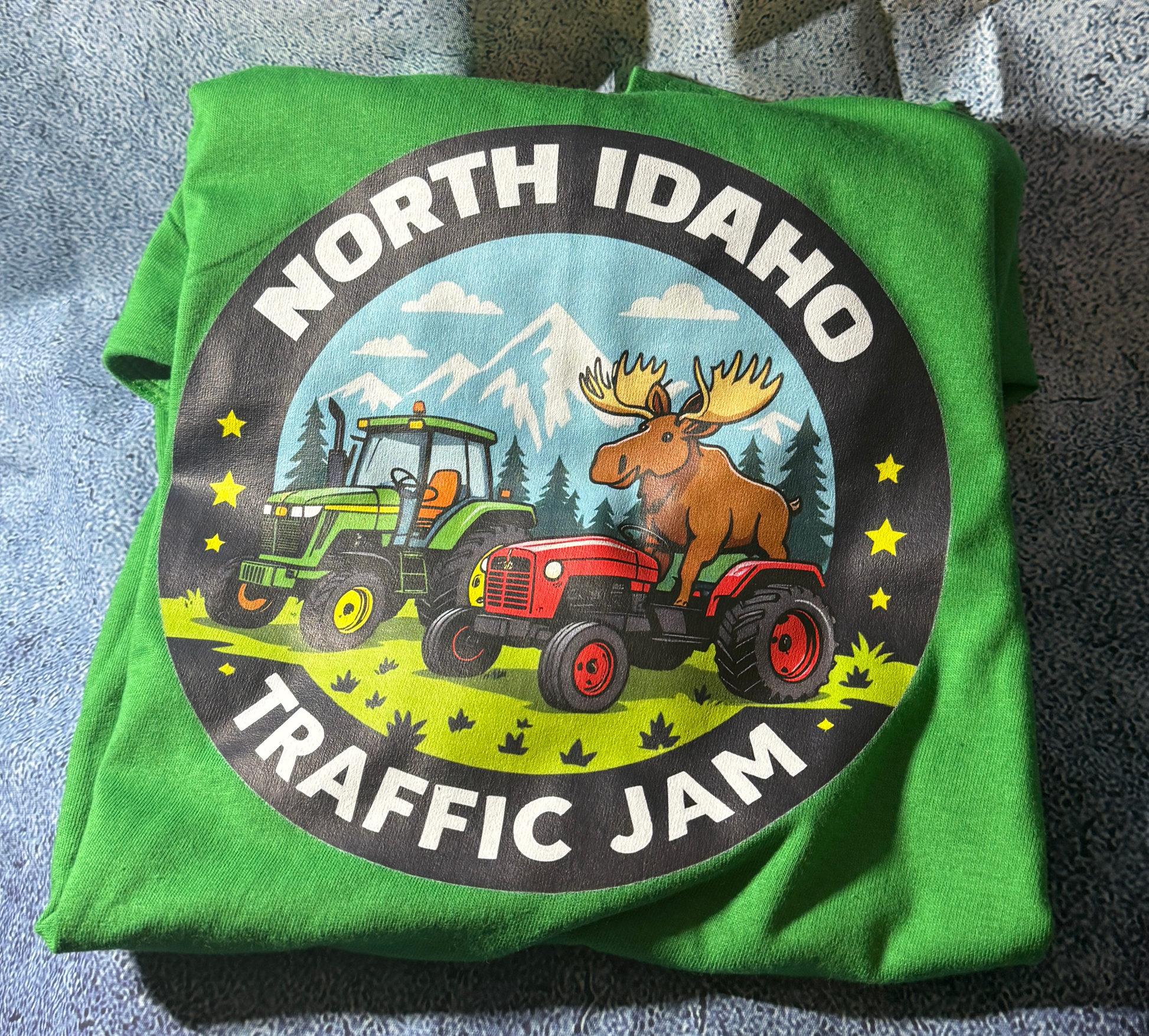 Green t-shirt with a graphic of a moose on a red tractor and text 'North Idaho Traffic Jam' on a gray surface.